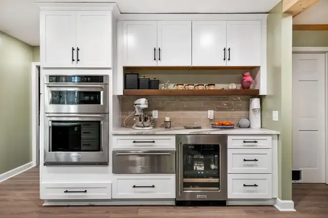 White kitchen wall with stacked wall ovens, a beverage fridge, open wood shelving and a tile backsplash